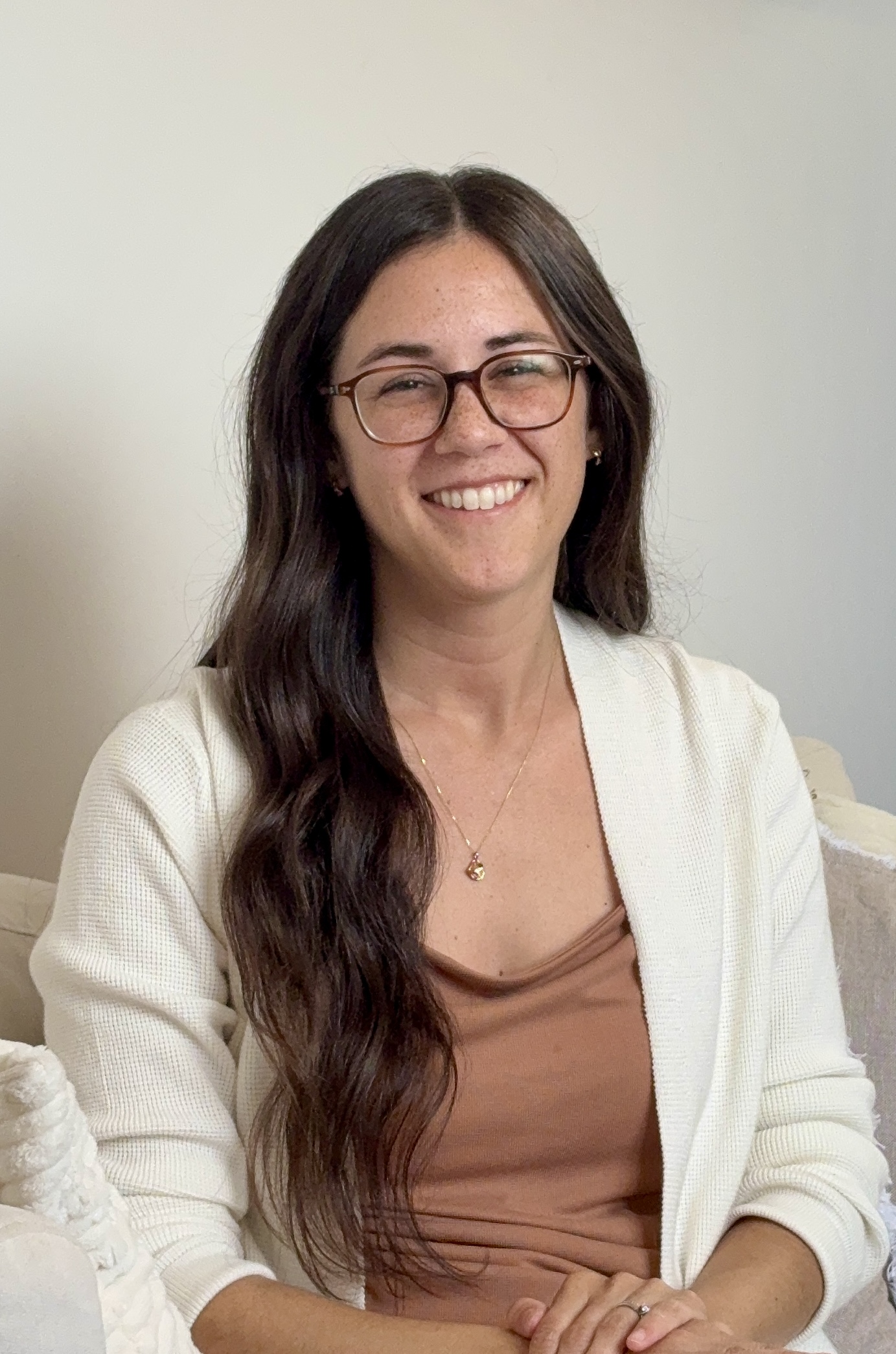 Megan Bugarin is wearing a white cardigan over a brown dress, she has long brown hair and glasses