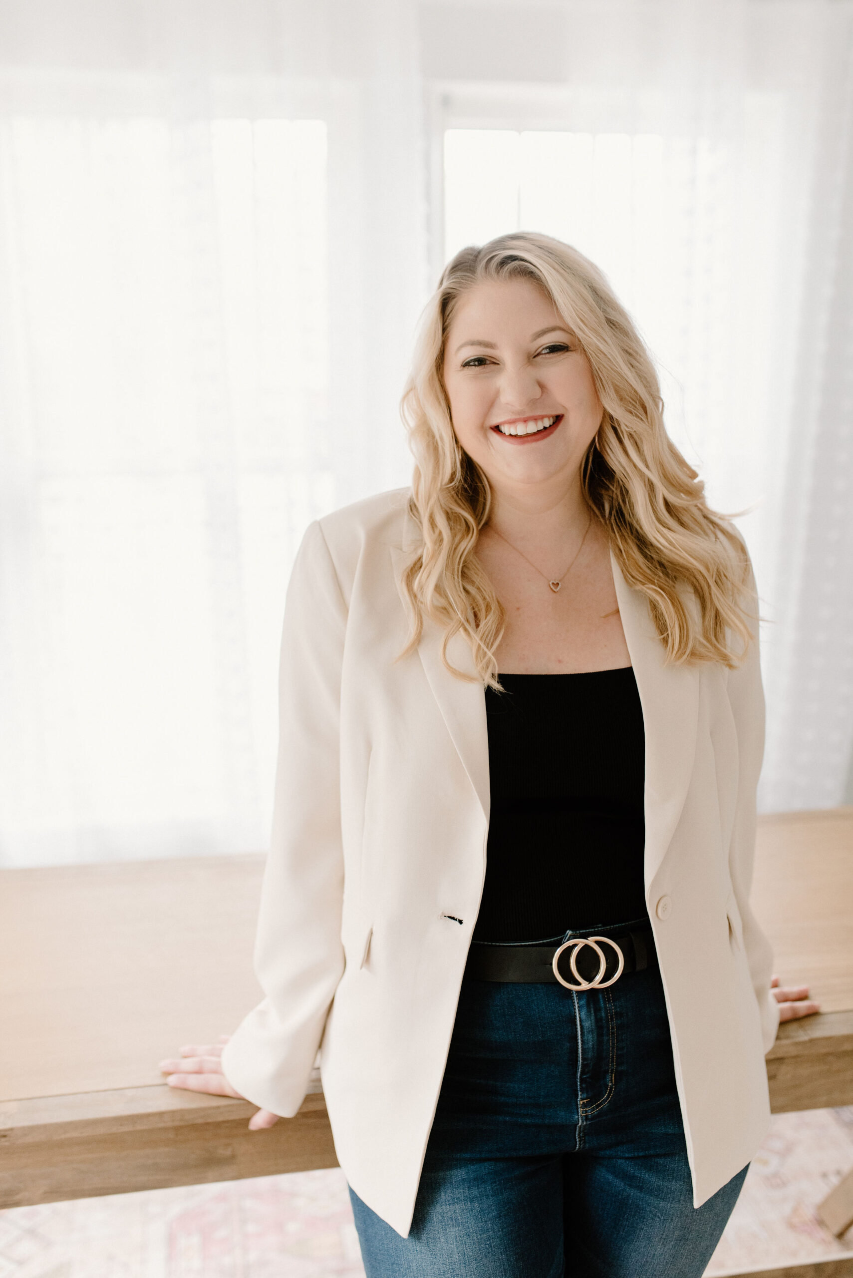 Rebecca, a white woman with blonde hair, wears a white blazer and learns against a table