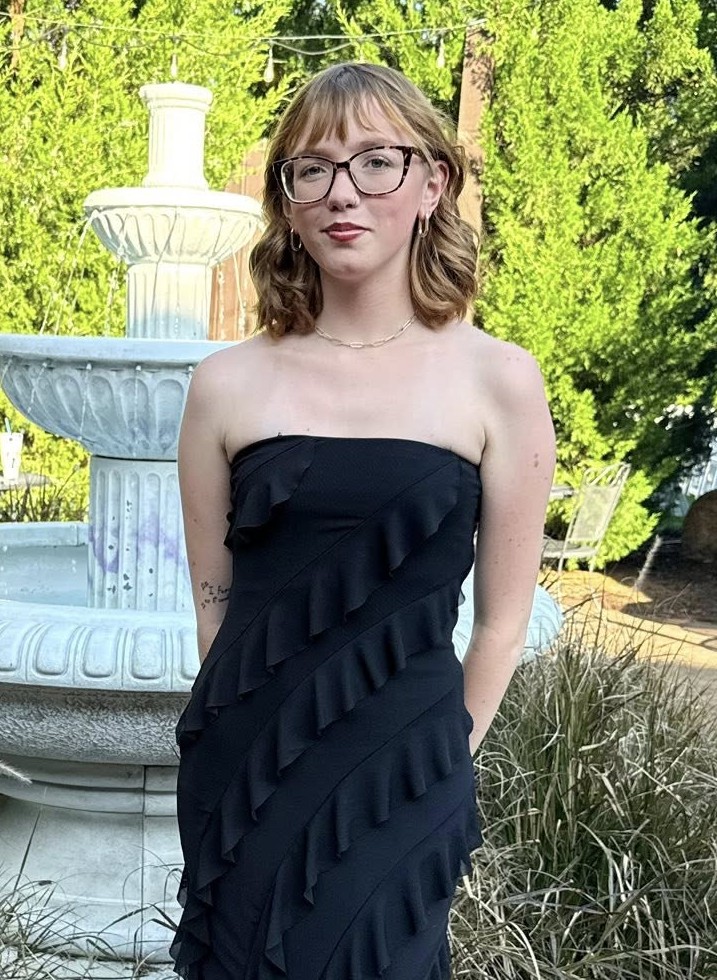 Stephanie, a white woman in a flowery shirt and grey dress pants, smiles at the camera while sitting in a wheelchair.